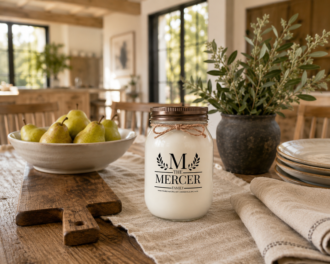 Customized Family Soy Candle personalized with a family name displayed on a reclaimed wood farmhouse dining table with fresh pears, neutral stoneware, linen napkins, olive branches, and handcrafted wood accents. The frosted white mason jar with distressed bronze lid creates a timeless family gathering scene filled with warmth and everyday traditions.