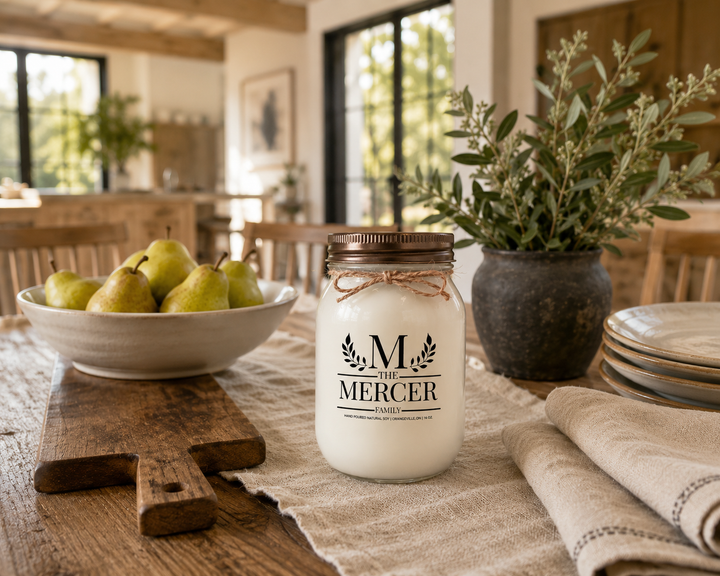Customized Family Soy Candle personalized with a family name displayed on a reclaimed wood farmhouse dining table with fresh pears, neutral stoneware, linen napkins, olive branches, and handcrafted wood accents. The frosted white mason jar with distressed bronze lid creates a timeless family gathering scene filled with warmth and everyday traditions.