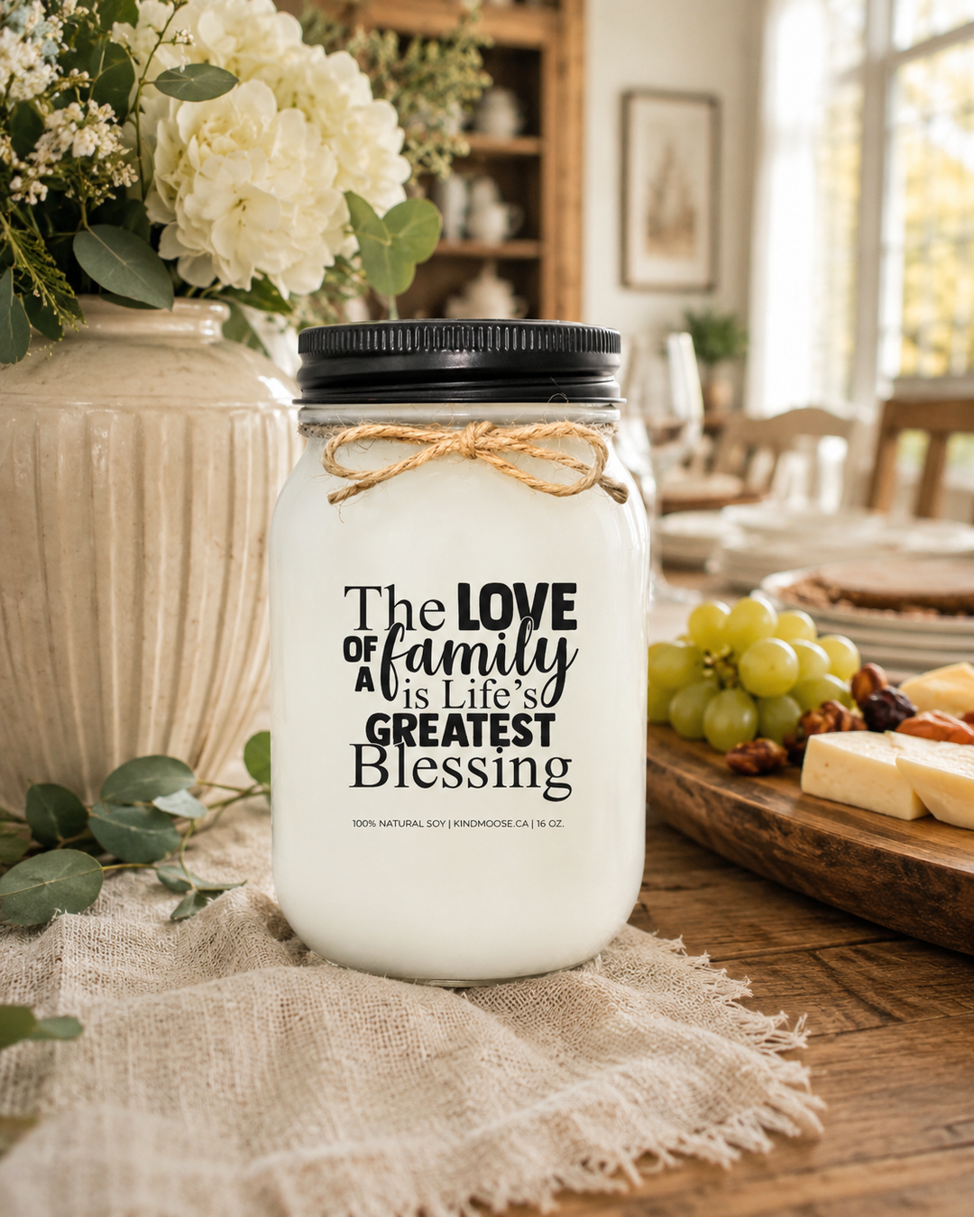 THE LOVE OF FAMILY IS LIFE'S GREATEST BLESSING soy candle displayed on a rustic farmhouse dining table surrounded by fresh flowers, greenery, warm wood textures, and elegant home décor. The welcoming family-inspired scene celebrates love, connection, gratitude, and the comfort of home.
