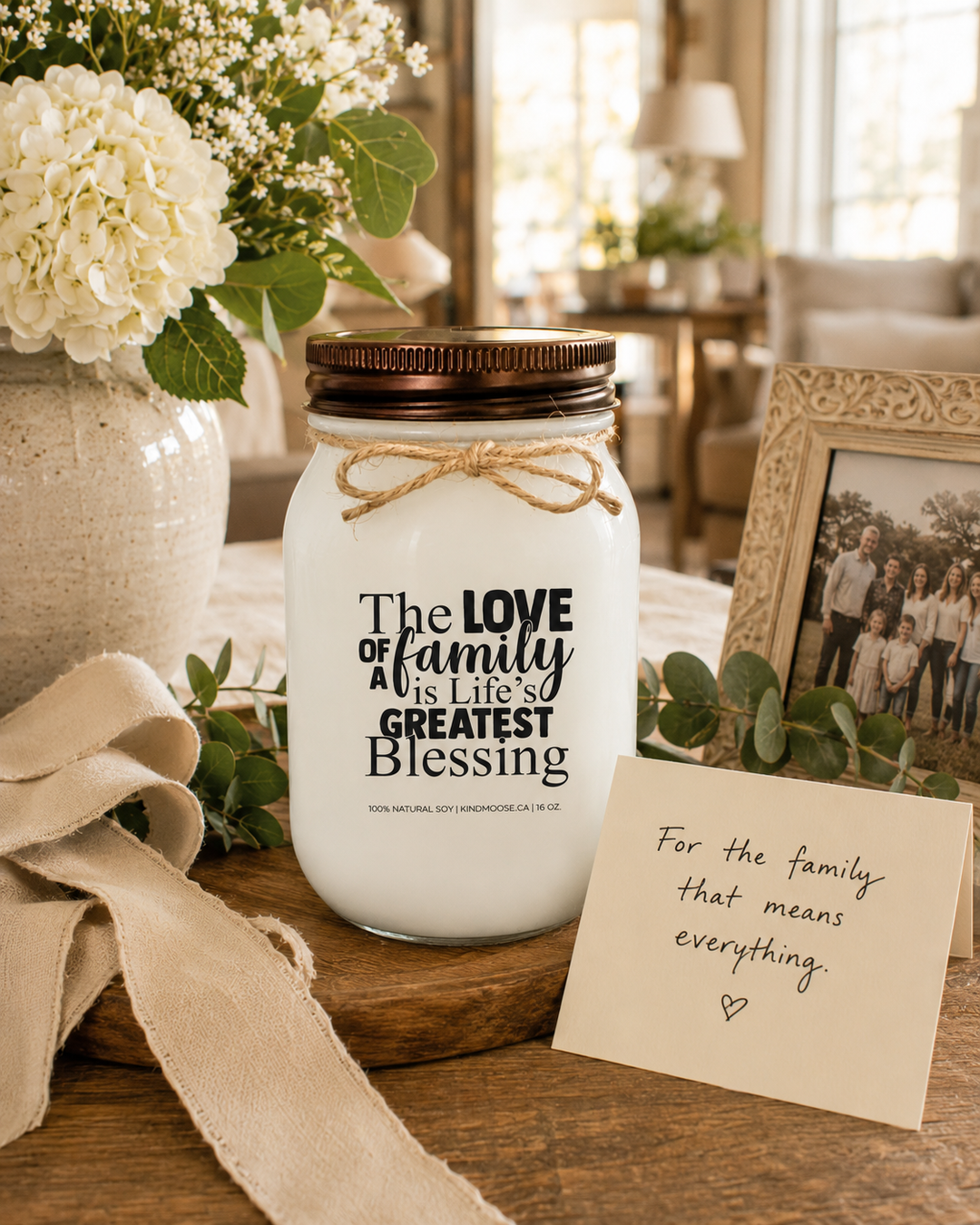 THE LOVE OF FAMILY IS LIFE'S GREATEST BLESSING soy candle with distressed bronze lid displayed as a thoughtful family gift on a farmhouse entry table. Surrounded by fresh flowers, a family photo frame, greenery, linen ribbon, and a handwritten gift card, the elegant gifting scene celebrates love, appreciation, and the importance of family.