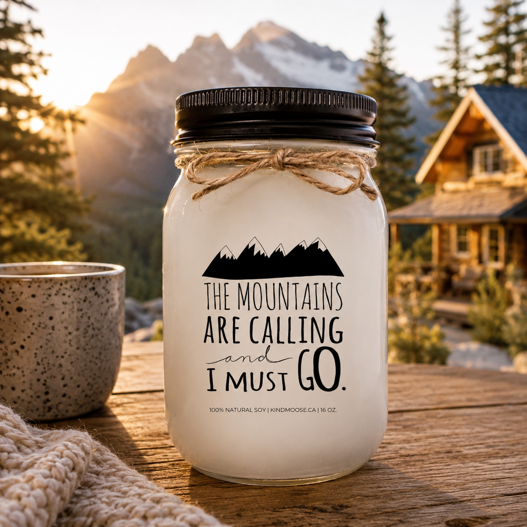 THE MOUNTAINS ARE CALLING AND I MUST GO soy candle with black lid displayed on a rustic mountain cabin table overlooking pine trees, mountain peaks, and a sunrise view.