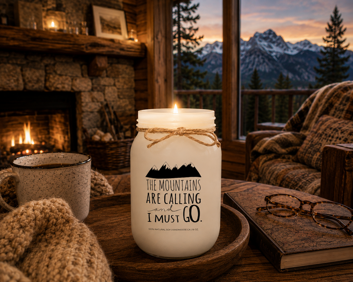 Burning THE MOUNTAINS ARE CALLING AND I MUST GO soy candle inside a cozy mountain cabin featuring a stone fireplace, mountain views, rustic furnishings, and warm evening lighting.