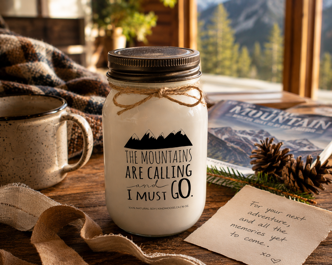 THE MOUNTAINS ARE CALLING AND I MUST GO soy candle with distressed bronze lid styled as a thoughtful mountain-inspired gift beside a handwritten card, travel magazine, coffee mug, pinecones, and evergreen sprigs.
