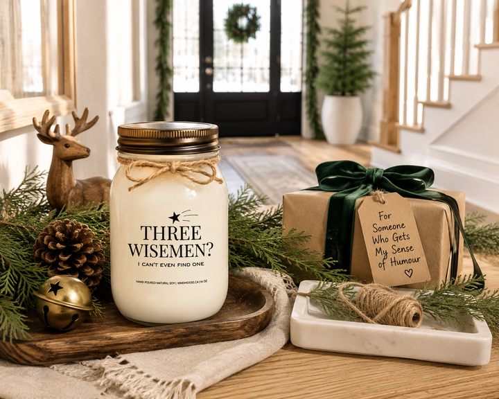 THREE WISE MEN? I CAN'T EVEN FIND ONE Christmas candle with a distressed bronze lid displayed on a reclaimed white oak entry console beside a beautifully wrapped gift, cedar greenery, pinecones, and antique brass accents in a bright luxury farmhouse foyer.
