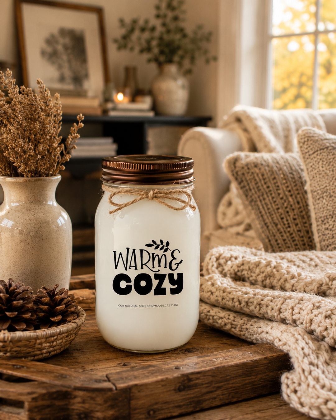 WARM & COZY soy candle with a distressed bronze lid styled in a bright farmhouse sunroom beside a rustic charcuterie board, artisan cheeses, crackers, grapes, herbal tea, and cozy knit textures. Warm afternoon sunlight and elegant neutral décor create a welcoming gathering space that celebrates slowing down, meaningful conversations, and comfortable living.