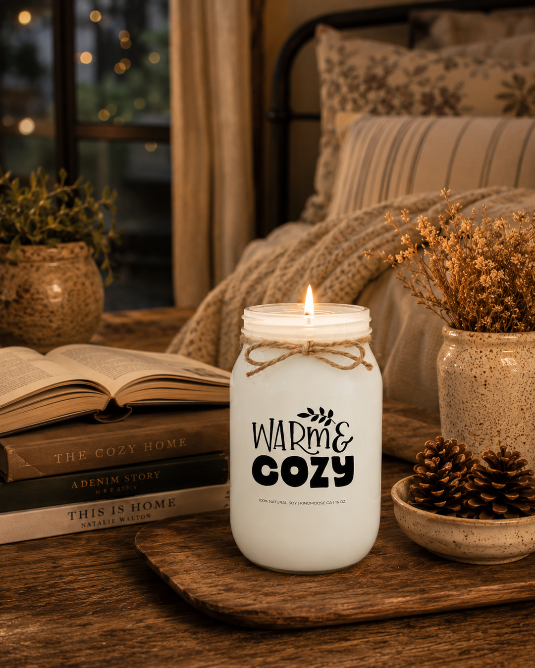 Burning WARM & COZY soy candle displayed on a rustic wooden side table in a peaceful farmhouse reading nook. Surrounded by an open novel, stacked books, a woven basket, pinecones, soft knit blankets, and warm ambient lighting, the cozy evening scene captures the comfort of quiet moments, relaxing at home, and enjoying a favorite book.