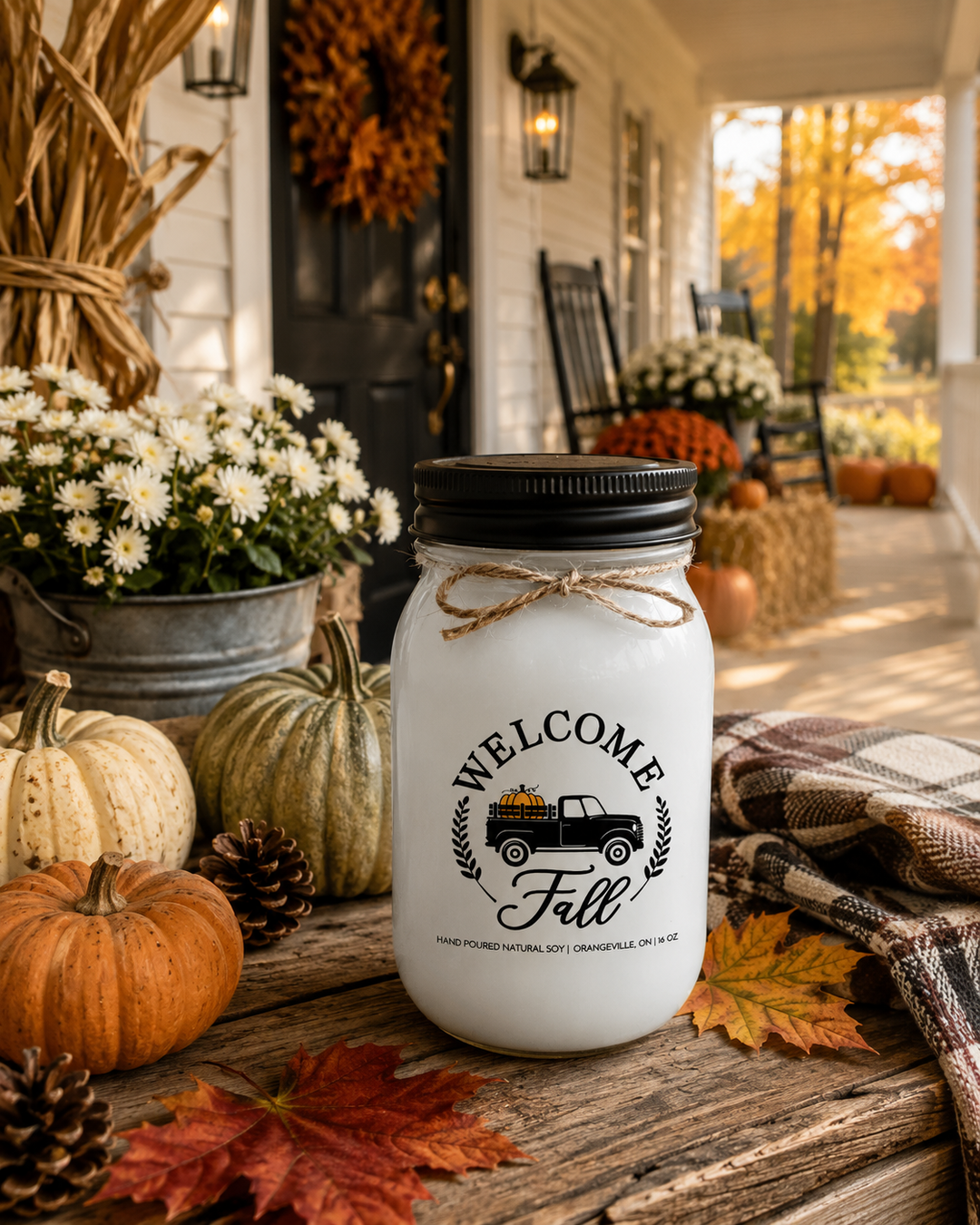 WELCOME FALL soy candle with a black lid displayed as the hero on a reclaimed wood farmhouse entry console surrounded by heirloom pumpkins, white mums, pinecones, colorful maple leaves, a rustic wooden crate, and elegant harvest décor. A charming white farmhouse porch with a black front door, autumn wreath, rocking chairs, and brilliant fall foliage creates a warm, welcoming entrance that celebrates the arrival of autumn.
