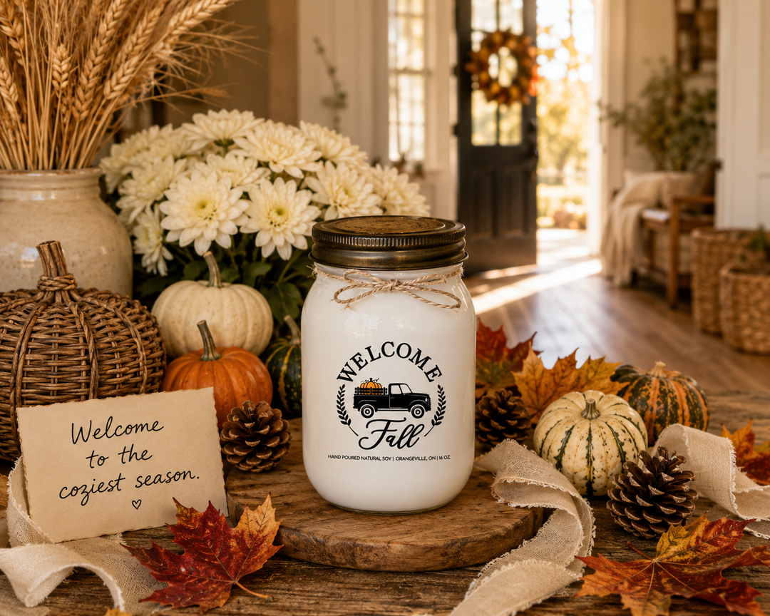 WELCOME FALL soy candle with a distressed bronze lid displayed as a thoughtful autumn gift on a rustic farmhouse entry console. Styled with a handwritten card reading “Welcome to the coziest season,” heirloom pumpkins, white chrysanthemums, colorful maple leaves, pinecones, a decorative wicker pumpkin, cream linen ribbon, and warm harvest textures. Golden afternoon sunlight fills the bright farmhouse foyer, creating a welcoming atmosphere that celebrates the beauty, warmth, and comfort of the autumn season.
