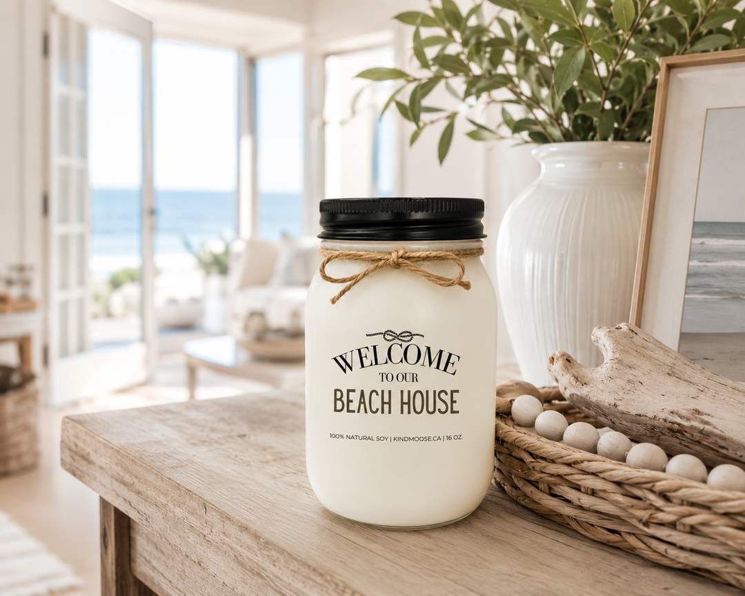 WELCOME TO OUR BEACH HOUSE soy candle with black lid displayed on a coastal entryway console table, surrounded by beach-inspired décor, greenery, driftwood accents, and bright ocean views in an elegant beach house.