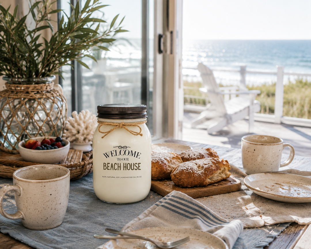 WELCOME TO OUR BEACH HOUSE soy candle with distressed bronze lid styled on a beach house breakfast table beside coffee mugs, fresh pastries, fruit, and coastal décor with ocean views through open patio doors.