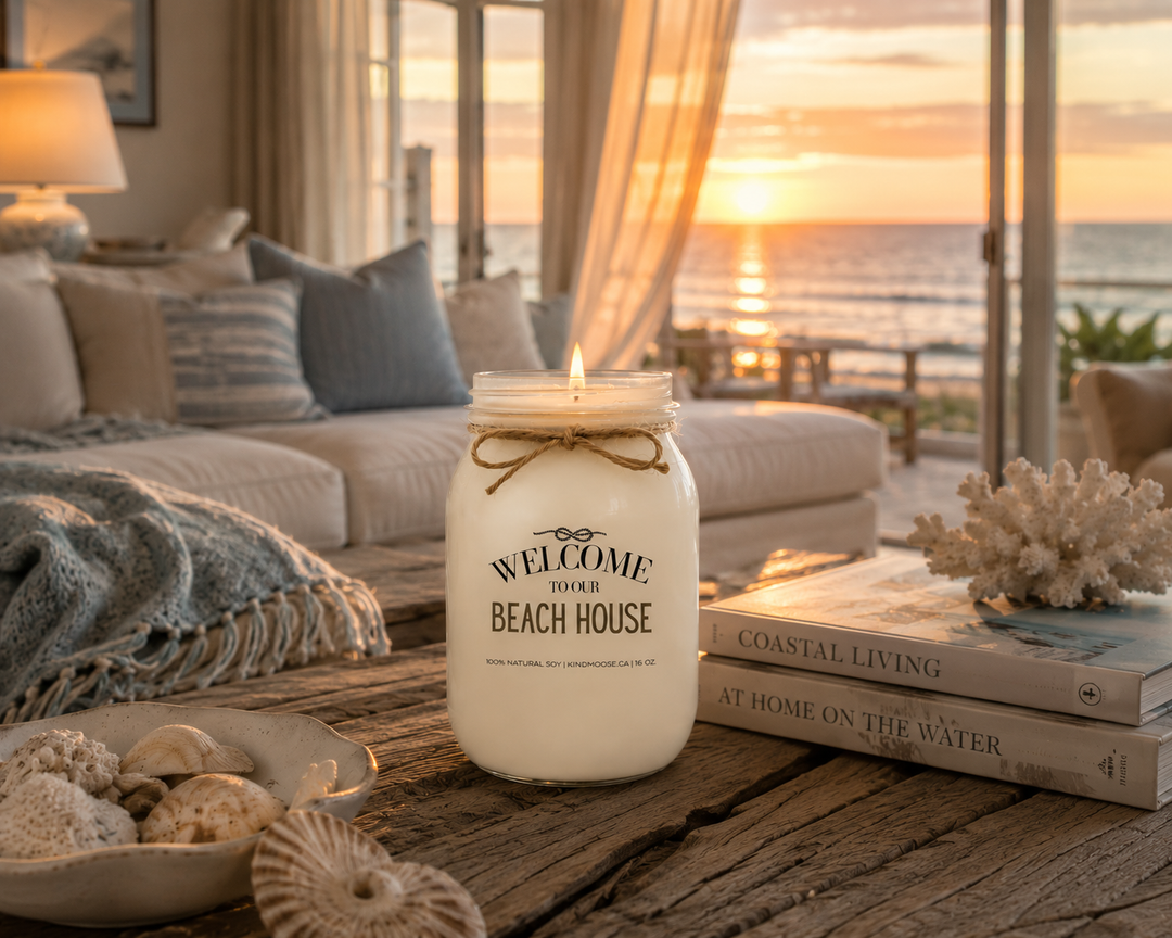 Burning WELCOME TO OUR BEACH HOUSE soy candle in a coastal living room at sunset, featuring a driftwood coffee table, ocean views, soft blue throws, coastal décor, and a peaceful beach house atmosphere.