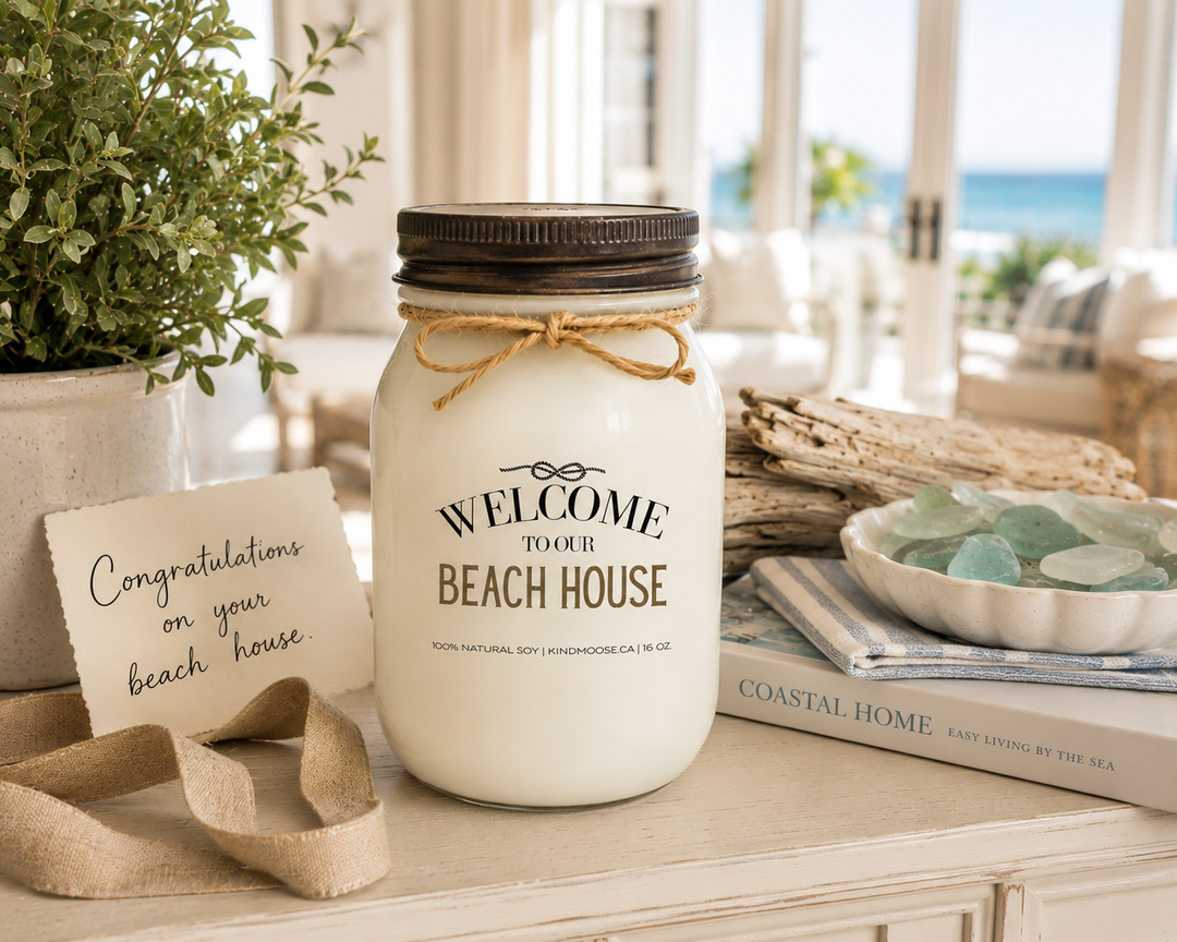 WELCOME TO OUR BEACH HOUSE soy candle with distressed bronze lid styled as a thoughtful beach house gift, featuring a handwritten congratulations card, linen ribbon, sea glass, driftwood, coastal greenery, and elegant beach-inspired décor.