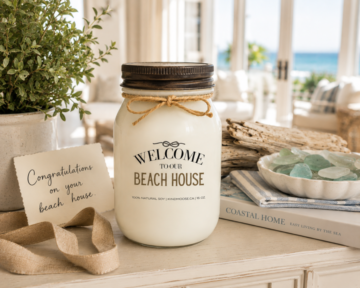 WELCOME TO OUR BEACH HOUSE soy candle with distressed bronze lid styled as a thoughtful beach house gift, featuring a handwritten congratulations card, linen ribbon, sea glass, driftwood, coastal greenery, and elegant beach-inspired décor.