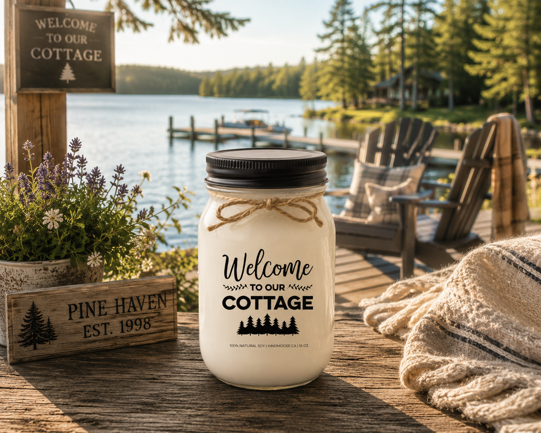 WELCOME TO OUR COTTAGE soy candle with black lid displayed on a rustic cottage deck overlooking a peaceful lake, surrounded by Adirondack chairs, dock views, cottage greenery, and warm morning sunlight.