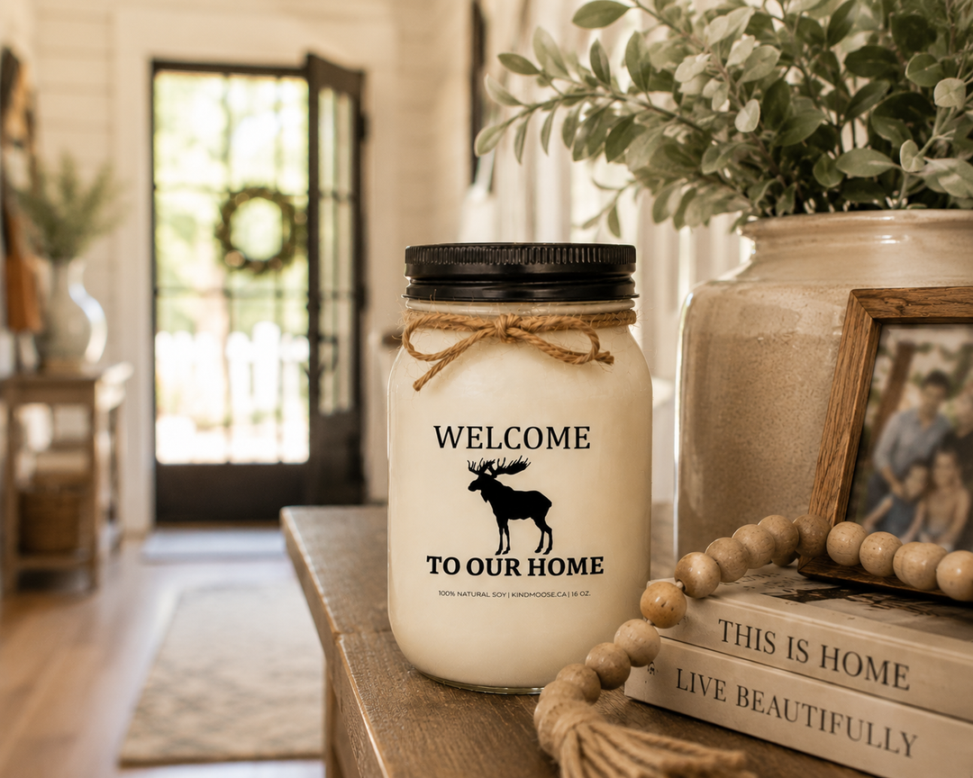 WELCOME TO OUR HOME soy candle with black lid displayed on an entryway console table in a bright modern farmhouse home, surrounded by greenery, family décor, and warm welcoming accents.
