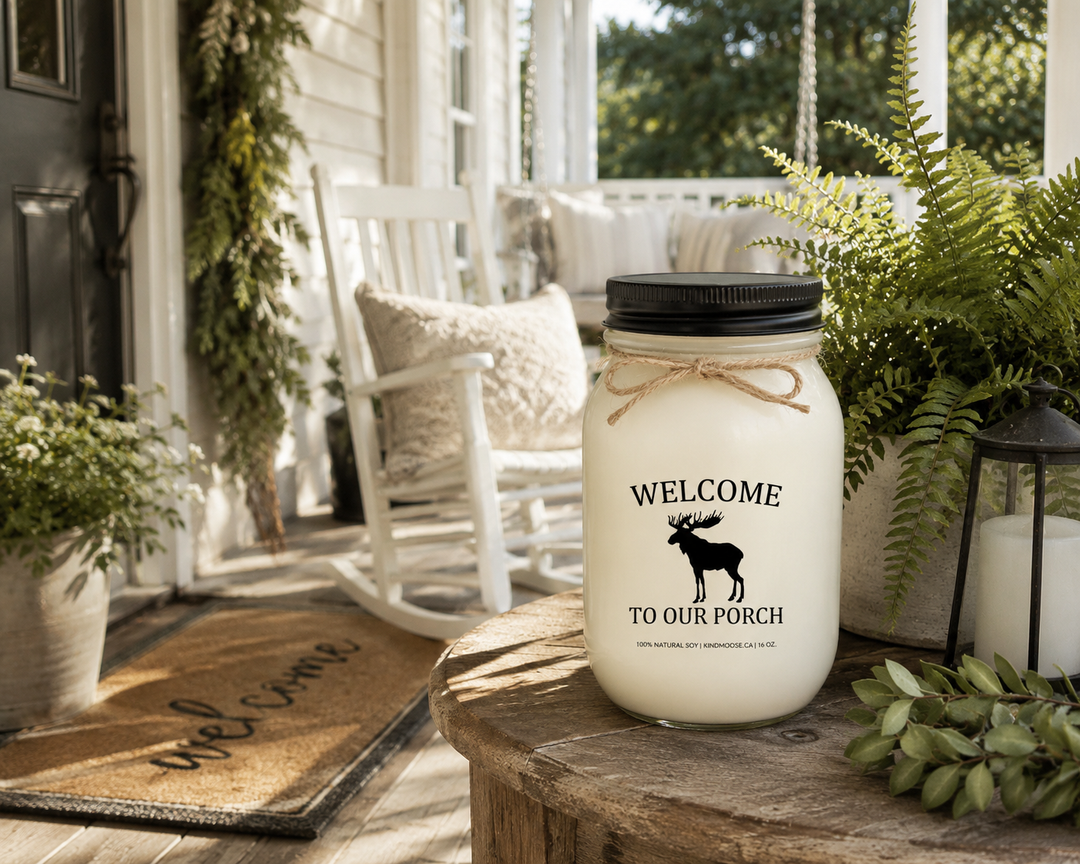 WELCOME TO OUR PORCH soy candle with black lid displayed on a welcoming farmhouse front porch beside a rocking chair, lantern, ferns, and rustic décor in warm morning sunlight.