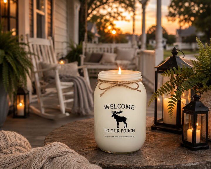 Burning WELCOME TO OUR PORCH soy candle on a farmhouse porch at sunset featuring rocking chairs, lanterns, ferns, soft blankets, and warm golden evening light.