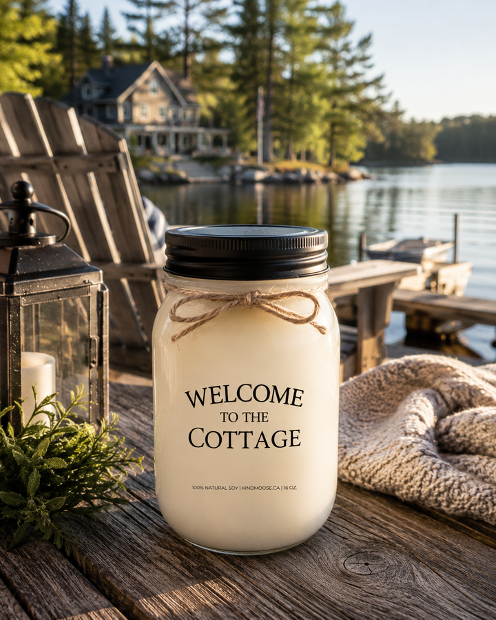 WELCOME TO THE COTTAGE soy candle with black lid displayed on a rustic cottage deck overlooking a peaceful lake, surrounded by Adirondack chairs, cottage greenery, and warm morning sunlight.