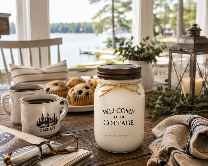 WELCOME TO THE COTTAGE soy candle with distressed bronze lid styled on a cottage breakfast table beside coffee mugs, blueberry muffins, reading glasses, and beautiful lake views through large windows.