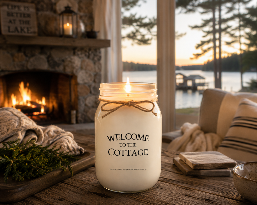 Burning WELCOME TO THE COTTAGE soy candle in a cozy lakeside cottage featuring a stone fireplace, soft blankets, rustic wood furnishings, and sunset views overlooking the lake.