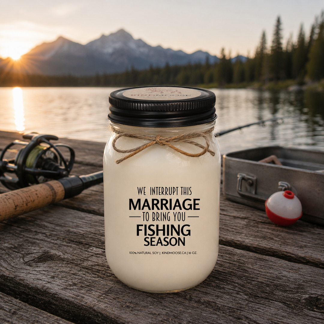 WE INTERRUPT THIS MARRIAGE TO BRING YOU FISHING SEASON soy candle with black lid displayed on a rustic cottage dock overlooking a peaceful lake at sunrise, surrounded by pine trees and calm morning water.