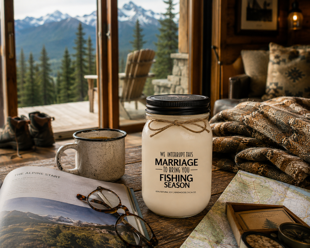 WE INTERRUPT THIS MARRIAGE TO BRING YOU FISHING SEASON soy candle with black lid displayed beside a fishing guidebook, reading glasses, coffee mug, lake map, wool blanket, and fishing boots inside a rustic cabin overlooking the outdoors.