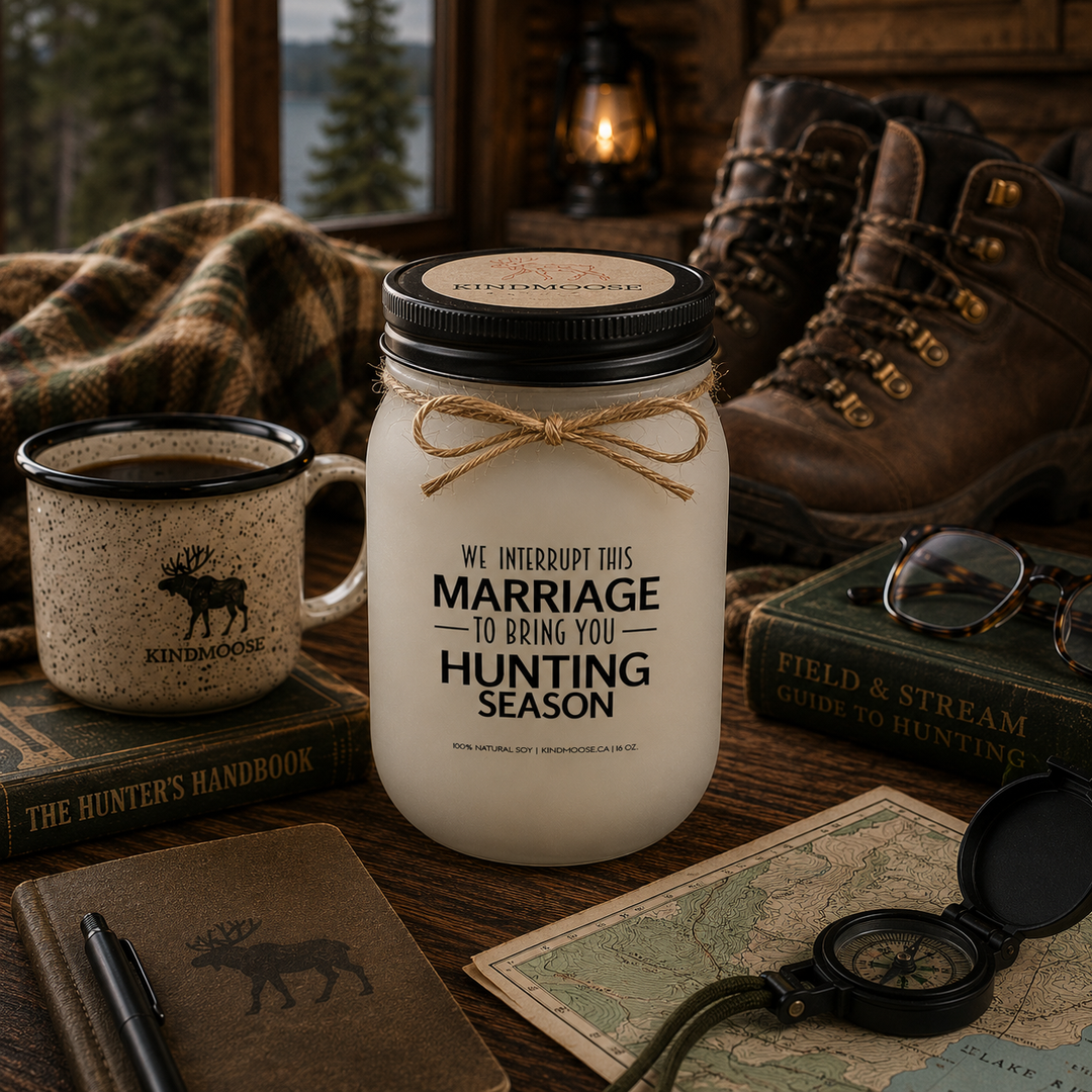 WE INTERRUPT THIS MARRIAGE TO BRING YOU HUNTING SEASON soy candle with black lid displayed beside a hunting guidebook, reading glasses, trail map, coffee mug, hiking boots, and wool blanket inside a warm mountain cabin.