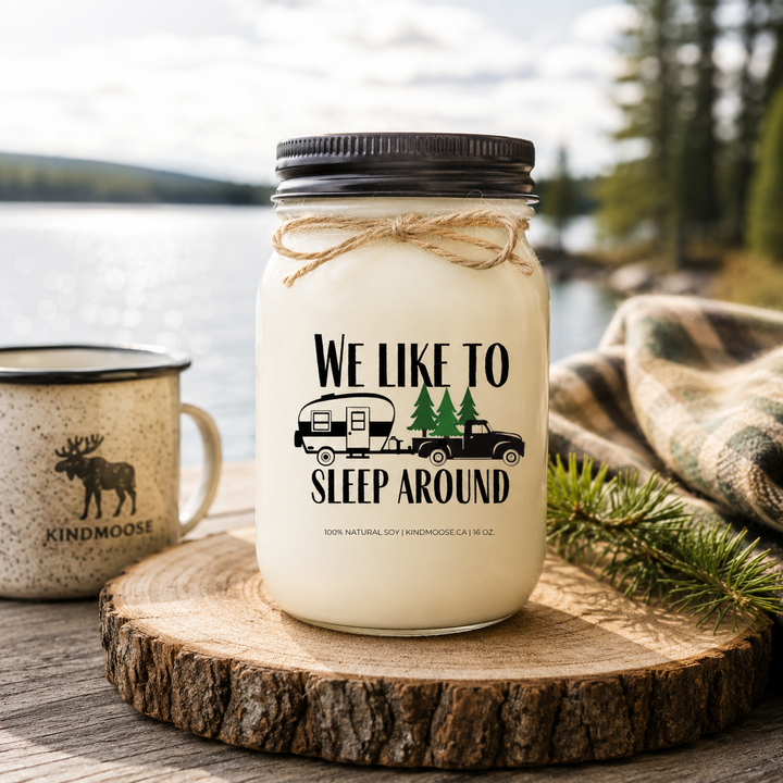 WE LIKE TO SLEEP AROUND soy candle with black lid displayed on a rustic campsite table overlooking a peaceful lake at sunrise, surrounded by pine trees, a vintage camper, and the beauty of outdoor adventure.