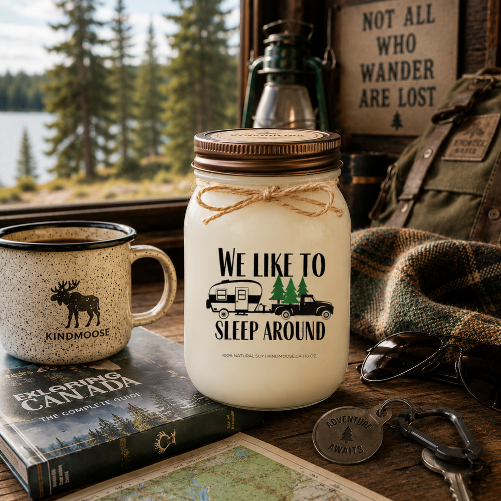 WE LIKE TO SLEEP AROUND soy candle with distressed bronze lid styled beside an enamel camping mug, campground guidebook, reading glasses, plaid blanket, and campsite map at a cozy RV campsite.