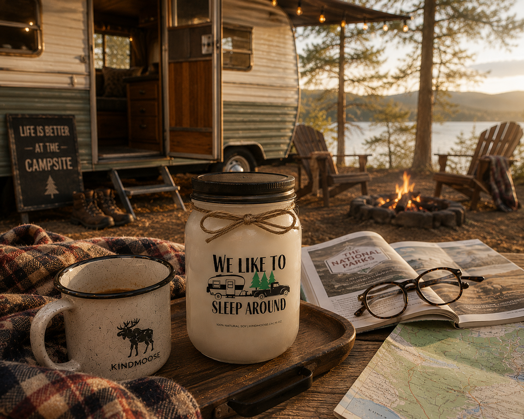 WE LIKE TO SLEEP AROUND soy candle with black lid displayed beside a campground guidebook, reading glasses, enamel coffee mug, plaid camping blanket, campground map, and hiking boots at a lakeside RV campsite.