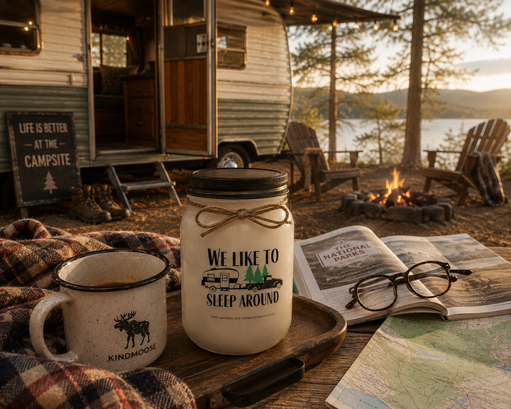 WE LIKE TO SLEEP AROUND soy candle with black lid displayed beside a campground guidebook, reading glasses, enamel coffee mug, plaid camping blanket, campground map, and hiking boots at a lakeside RV campsite.