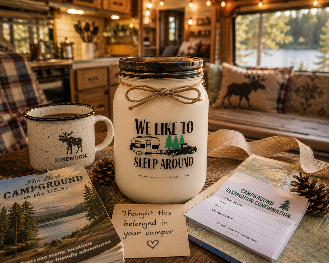 WE LIKE TO SLEEP AROUND soy candle with distressed bronze lid styled as a thoughtful camping gift inside a cozy camper, featuring a handwritten note, campground reservation confirmation, camping magazine, enamel mug, pinecones, and rustic RV décor.