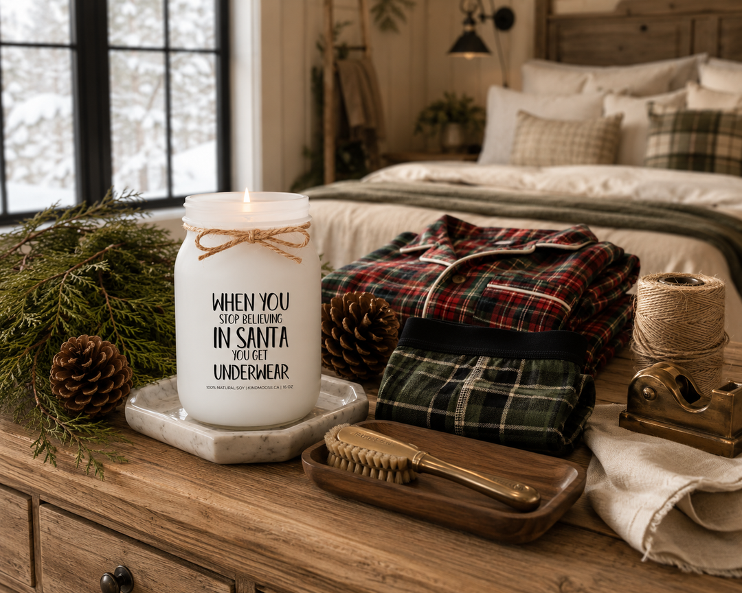 WHEN YOU STOP BELIEVING IN SANTA YOU GET UNDERWEAR customizable Christmas candle burning on a reclaimed white oak bedroom dresser beside folded plaid Christmas pajamas, premium underwear, cedar sprigs, pinecones, and walnut accessories inside a peaceful luxury Canadian farmhouse bedroom.