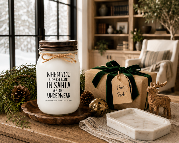 WHEN YOU STOP BELIEVING IN SANTA YOU GET UNDERWEAR customizable Christmas candle with a distressed bronze lid displayed beside a beautifully wrapped Christmas gift, cedar branches, pinecones, a brass sleigh bell, and a carved wooden reindeer on a reclaimed white oak library console inside a luxury Canadian farmhouse.