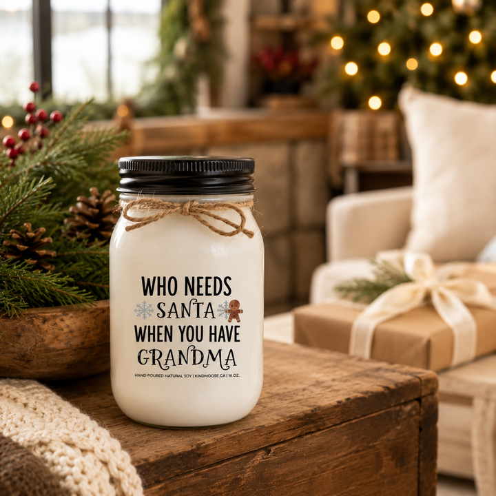 KINDMOOSE "Who Needs Santa When You Have Grandma" 16 oz frosted white soy candle with an antique gold lid displayed on a rustic farmhouse entry table decorated with cedar garland, Christmas wreath, stockings and wrapped gifts in warm holiday sunlight.