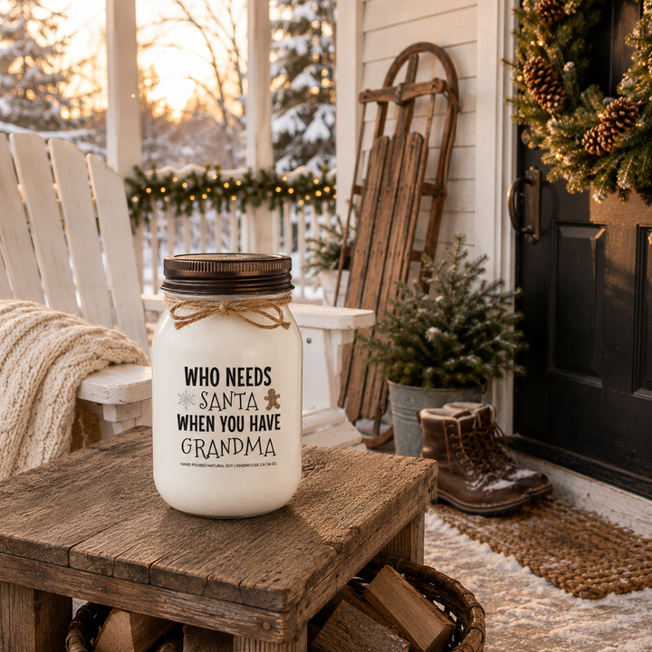 KINDMOOSE "Who Needs Santa When You Have Grandma" 16 oz frosted white soy candle with a signature distressed bronze lid on a snow-covered farmhouse porch beside an Adirondack chair, cedar garland and winter décor in warm golden afternoon light.