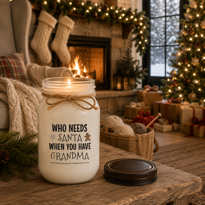 KINDMOOSE "Who Needs Santa When You Have Grandma" 16 oz frosted white soy candle burning with a natural flame on a rustic farmhouse side table, distressed bronze lid beside the candle, surrounded by a cozy fireplace, Christmas tree and festive holiday décor.