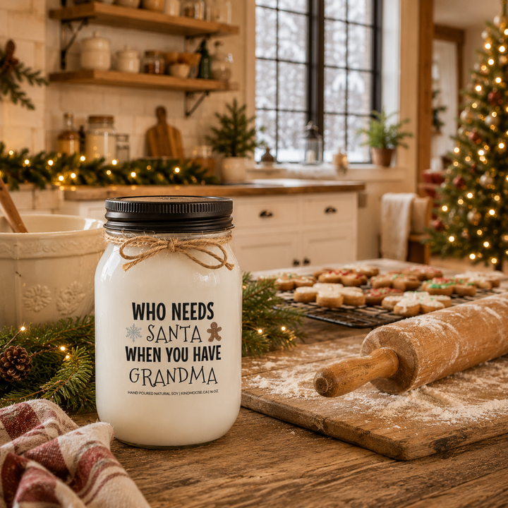 KINDMOOSE "Who Needs Santa When You Have Grandma" 16 oz frosted white soy candle with a black lid displayed on a farmhouse kitchen island beside a rolling pin, fresh Christmas cookies and holiday baking décor, inspired by treasured Christmas traditions with Grandma.