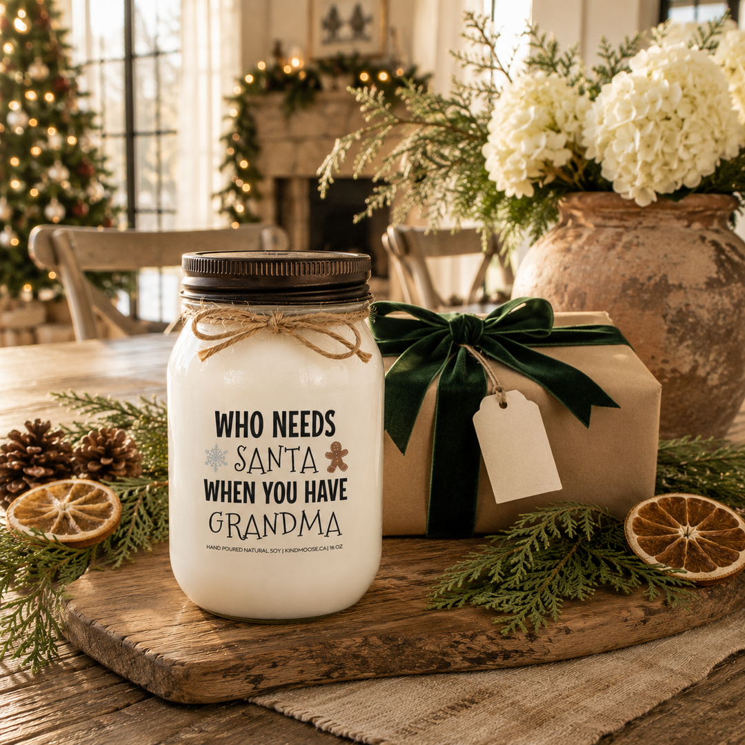 KINDMOOSE "Who Needs Santa When You Have Grandma" 16 oz frosted white soy candle with a signature distressed bronze lid presented as a thoughtful Christmas gift beside a wrapped present, cedar garland, pinecones and white hydrangeas in a cozy farmhouse dining room.